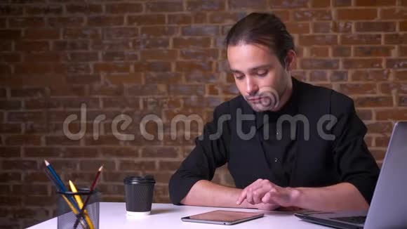 年轻的白种人商人一边在电脑旁坐着一边在桌旁挥舞着他的设备一边喝着咖啡视频的预览图