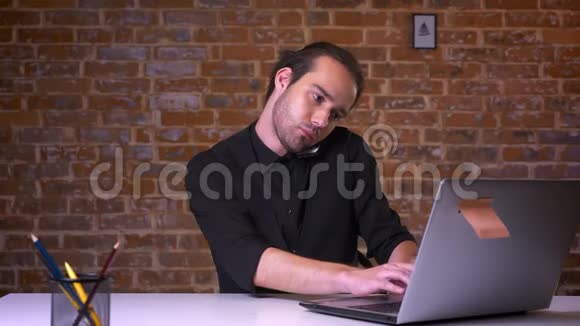 帅气的年轻人坐在砖头上一边看电脑一边拿着电话一边等着接视频的预览图