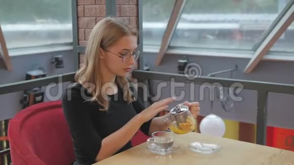 一个戴着眼镜的小女孩坐在咖啡馆里把茶从茶壶里倒进杯子里视频的预览图