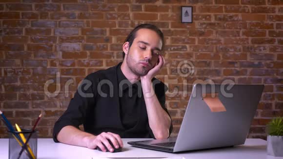 白种人的商人一边打哈欠一边看着笔记本电脑一边坐在书桌旁轻松地带着砖背景视频的预览图