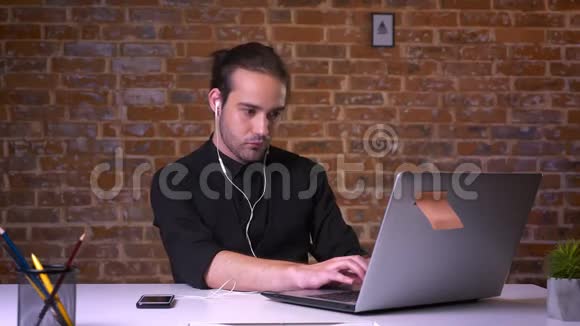 在工作室里戴着耳机听音乐在砖墙旁的笔记本电脑上打字的可爱白种人放松了视频的预览图