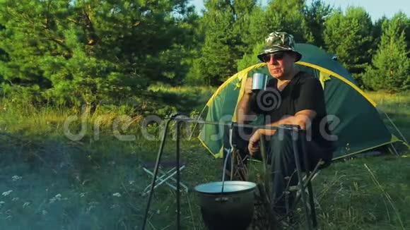一个戴墨镜的人坐在帐篷附近的火堆上喝茶欣赏大自然视频的预览图