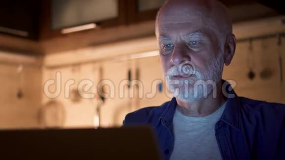 老人在家里的办公室里晚上使用笔记本电脑时戴上无线耳机听音乐视频的预览图