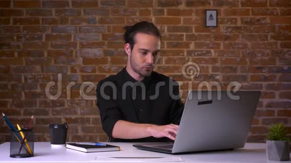 有魅力的白种人年轻男性喝着咖啡在红工作室里的工作场所看着电脑视频的预览图