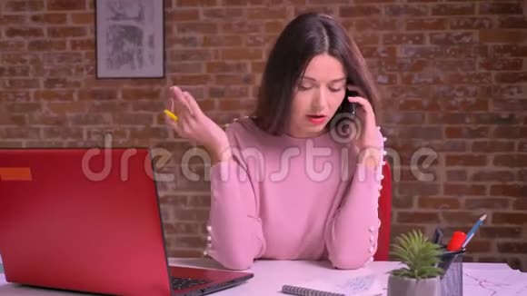 可爱的白种人女性在笔记本电脑前自信而专注地在手机上讨论她的生意视频的预览图
