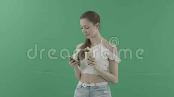 迷人的女人在绿色背景下喝鸡尾酒在智能手机上发短信视频的预览图