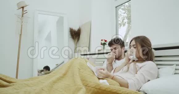 一对生病的年轻夫妇流鼻涕他们在温暖的毯子下躺在床上喝茶视频的预览图