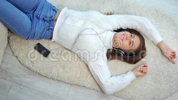 积极的年轻女性躺在蓬松的地毯上用智能手机和耳机听音乐视频的预览图
