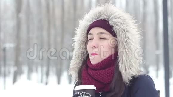 美丽的年轻女孩呼出寒冷的空气微笑和喝咖啡从一个杯子在冬季公园视频的预览图
