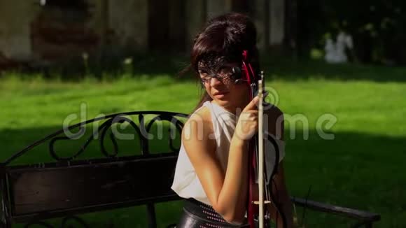 美丽的年轻女孩坐在长凳上在美丽的公园里拉小提琴视频的预览图