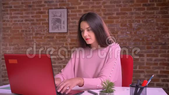 白色快乐的女商人坐在桌子旁穿着粉色毛衣在笔记本电脑上工作打字视频的预览图