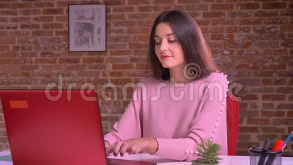 成年白种人女性在笔记本电脑上打字坐在桌子旁的砖背景下做梦视频的预览图