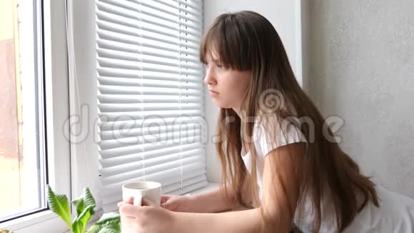 十几岁的女孩在靠近窗户的窗台上喝茶美丽悲伤的女孩望着窗外喝茶或咖啡视频的预览图
