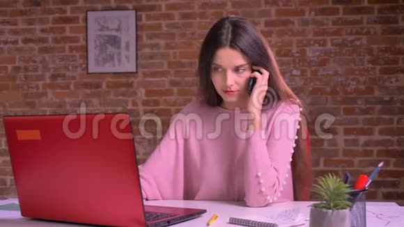 漂亮的年轻商务女性穿着粉红色毛衣在打电话看着她的笔记本电脑说话视频的预览图