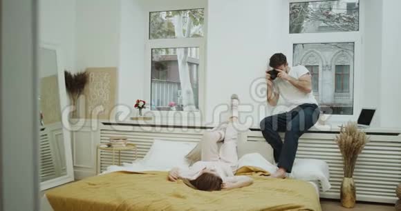 年轻人在他们舒适的卧室里给女朋友拍照她躺在床上穿着一件可爱的粉红色衣服视频的预览图