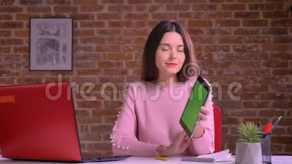 漂亮的白种人商务女性展示着绿色的屏幕在砖墙旁边的桌子旁开心而轻松地微笑着视频的预览图