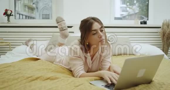 有魅力的年轻女人在她的笔记本上敲击她躺在床上穿着一件粉红色的时尚睡衣视频的预览图