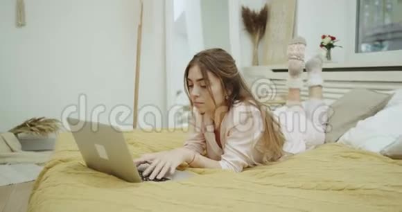 一位穿睡衣的可爱女士晚上在她的现代卧室里花时间在床上的笔记本上视频的预览图
