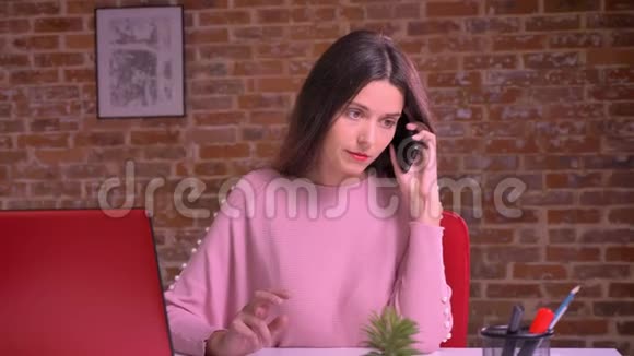 年轻漂亮的白种人女孩坐在红色工作室的工作场所一边打着电话视频的预览图