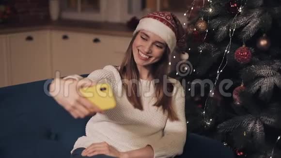 戴着圣诞帽坐在沙发上的年轻可爱的快乐女孩在圣诞树旁做自我选择视频的预览图