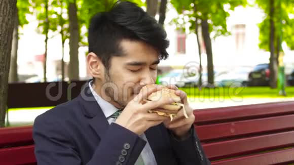 亚洲商人坐在公园的长椅上吃着美味的大汉堡视频的预览图
