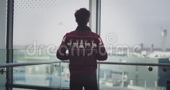 一个年轻人在一个现代化的建筑旁边的大窗户站着背对着镜头穿着红色的毛衣享受着视频的预览图