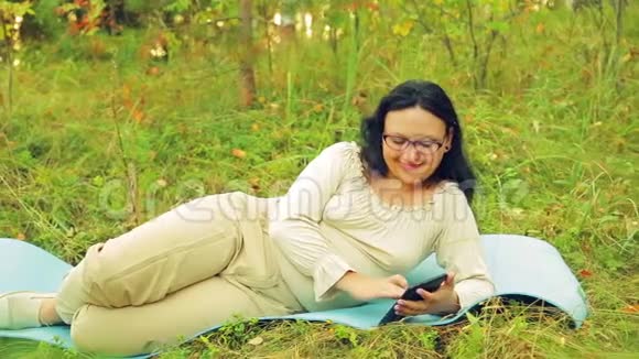 森林里一个戴眼镜的年轻微笑的女人躺在地毯上做瑜伽并在信使中交流视频的预览图