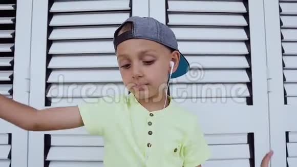 一个穿着t恤和帽子的可爱男孩的肖像他用耳机在智能手机上听最喜欢的音乐跳舞视频的预览图