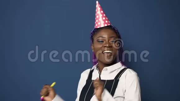 快乐的非洲女人戴着生日帽跳舞在蓝色背景上吹着生日喇叭视频的预览图
