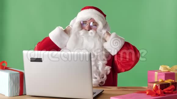 圣诞老人微笑着坐在笔记本电脑后面听着耳机上的音乐和跳舞背景是绿色的色度视频的预览图