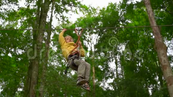 一个小男孩在森林探险公园的树梢上的高空滑索上穿着安全装备的慢速镜头室外视频的预览图
