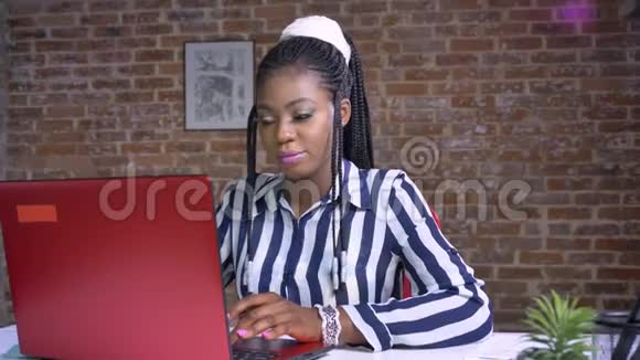 漂亮的非洲女性看着她的电脑然后看着相机开心和放松坐在砖头办公室视频的预览图