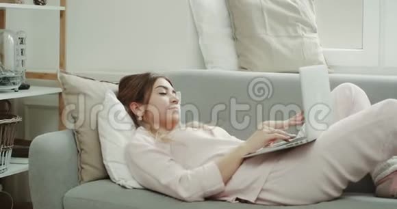 现代设计在客厅年轻的女人敲击笔记本她正穿着睡衣坐在沙发上视频的预览图