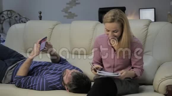 沙发上的金发妻子在笔记本上读笔记丈夫躺在附近在电话旁聊天视频的预览图