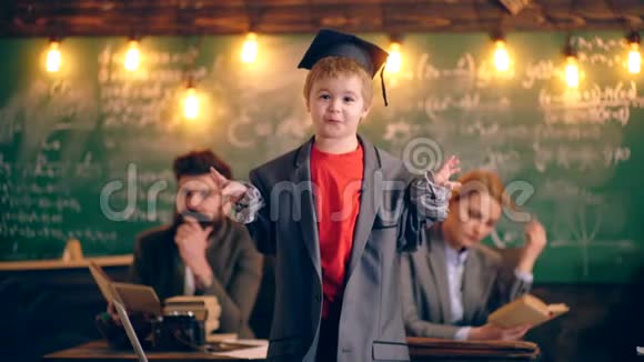 穿着成人夹克和毕业帽的男孩在学校教室里以老师为背景讲述故事视频的预览图