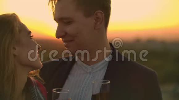 男人和年轻漂亮的女人坐在沙发上拿着酒杯在日落时亲吻屋顶露台视频的预览图