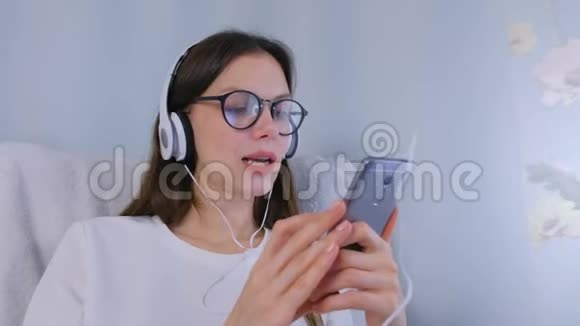 戴眼镜的快乐黑发女人正在智能手机上用耳机听音乐唱一首歌视频的预览图