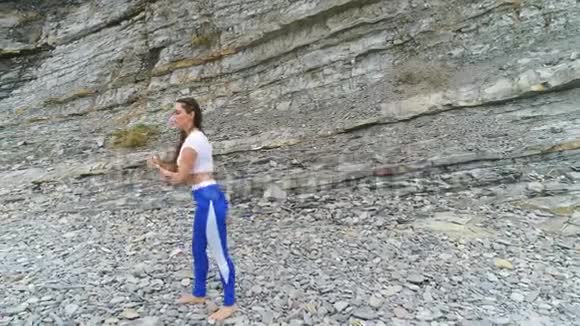女孩正在做腿部运动在海洋和岩石背景上的呼吸练习中做身体反射视频的预览图