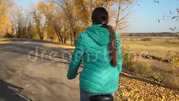 穿绿色夹克的年轻女孩骑自行车在秋天公园的道路上在黄树的背景下视频的预览图