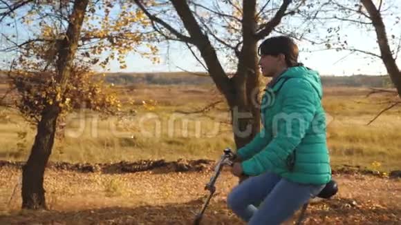 穿绿色夹克的年轻女孩骑自行车在秋天公园的黄色树木背景上慢动作视频的预览图