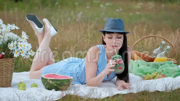 一个戴着帽子穿着衣服的年轻美女坐在草地上的格子花格子上喝着罐装柠檬水视频的预览图