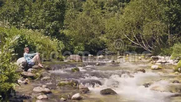 夏季徒步旅行时在石河岸边放松的女人野生自然景观视频的预览图