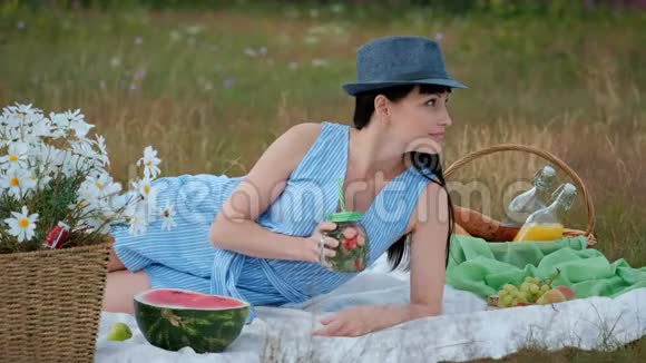 一个戴着帽子穿着衣服的年轻美女坐在草地上的格子花格子上喝着罐装柠檬水视频的预览图