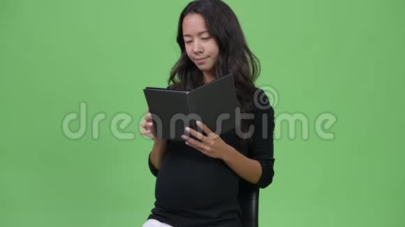 年轻美丽的怀孕多民族妇女一边读书一边摸肚子视频的预览图