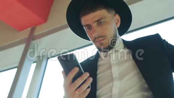 穿着西装戴着一顶带胡须的帽子的时尚白种人男性商人站在智能手机上阅读信息视频的预览图