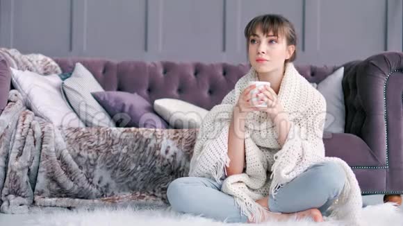 年轻女子裹着柔软蓬松的格子布端着杯子坐在家里的白色地毯上视频的预览图