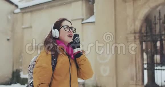 白雪皑皑的城市里漂亮迷人的高加索穿洞女孩她正在用大白耳机听音乐在路上行走视频的预览图