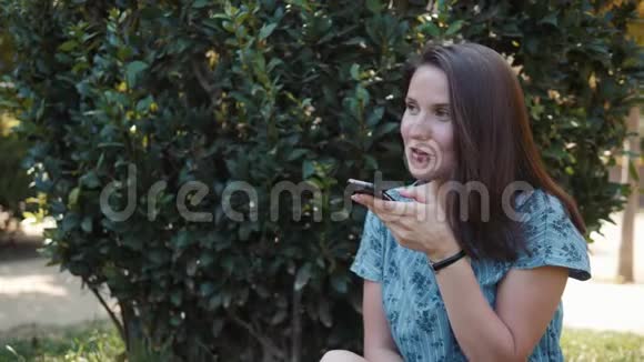 快乐的年轻女孩黑发女孩夏天坐在公园的户外使用手机与语音识别ai音频视频的预览图