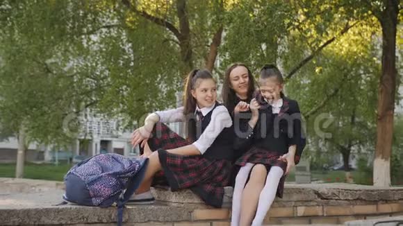 母亲带着两个穿校服的女儿背包坐在学校附近的街道上亲吻和交谈视频的预览图