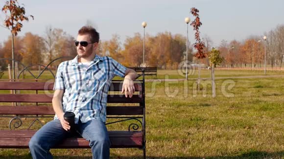 一个戴着黑眼镜的年轻白种人坐在公园里的长凳上喝着咖啡慢节奏复制空间视频的预览图
